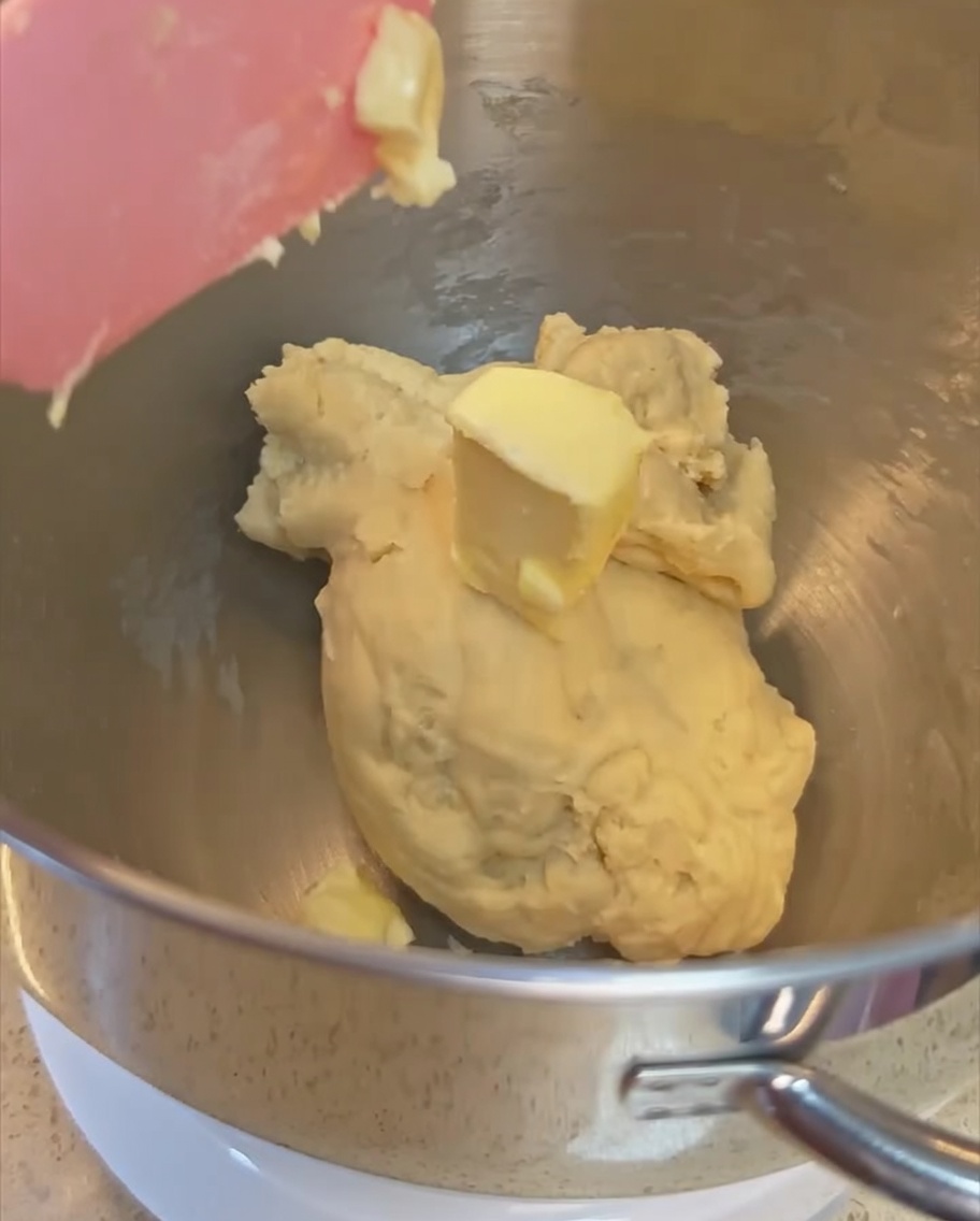 Un trozo de mantequilla siendo añadido a la masa parcialmente amasada dentro del bol de acero inoxidable.