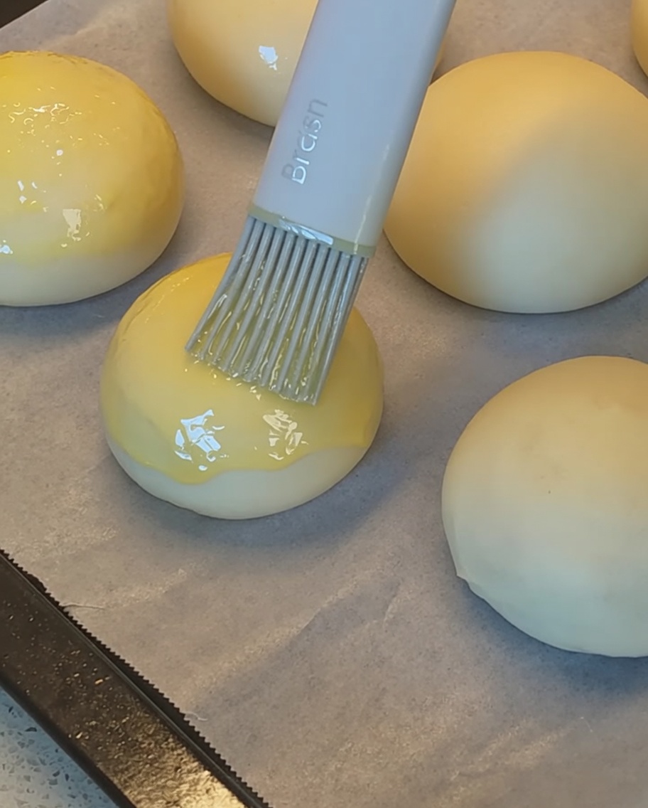 Un pincel de silicona aplicando huevo batido sobre las piezas de masa antes de espolvorear semillas de sésamo.