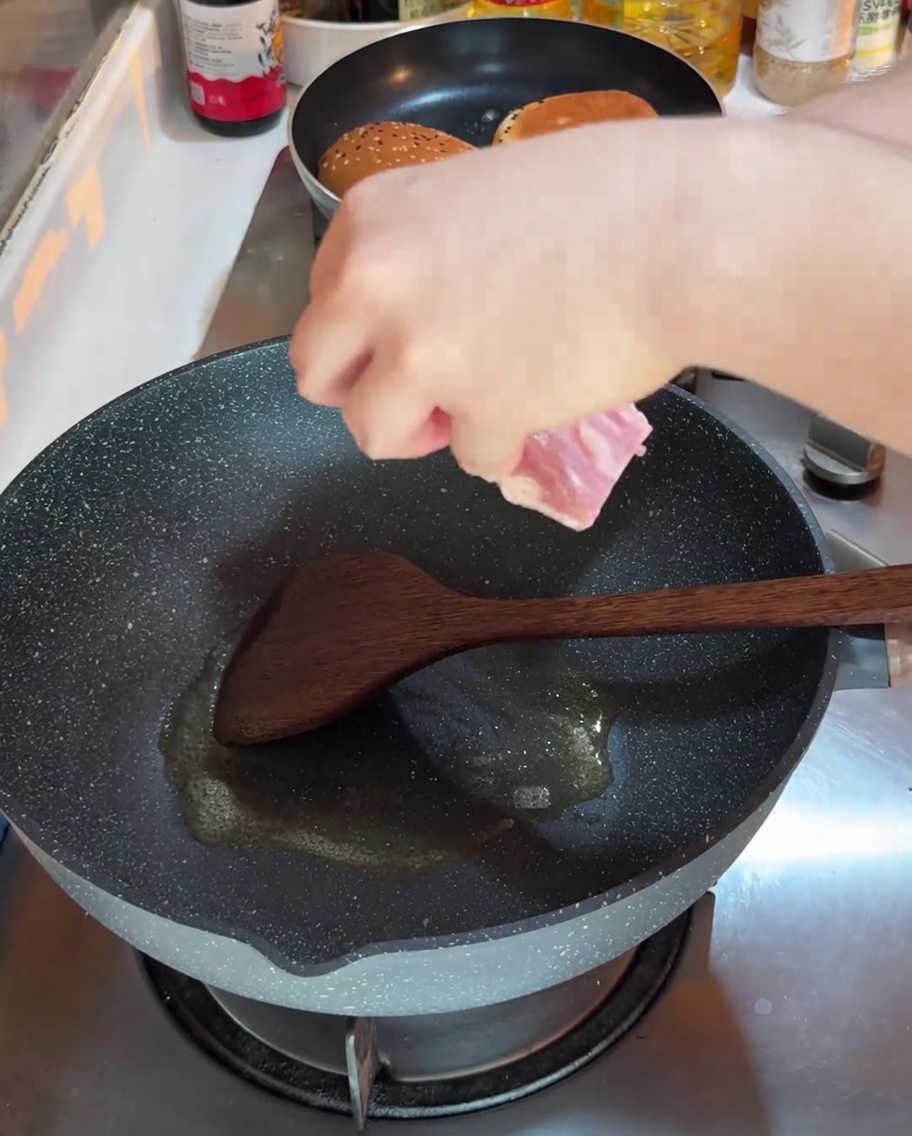 Una mano colocando una tira de bacon crudo en un wok caliente para freír, con una sartén más pequeña visible al fondo.