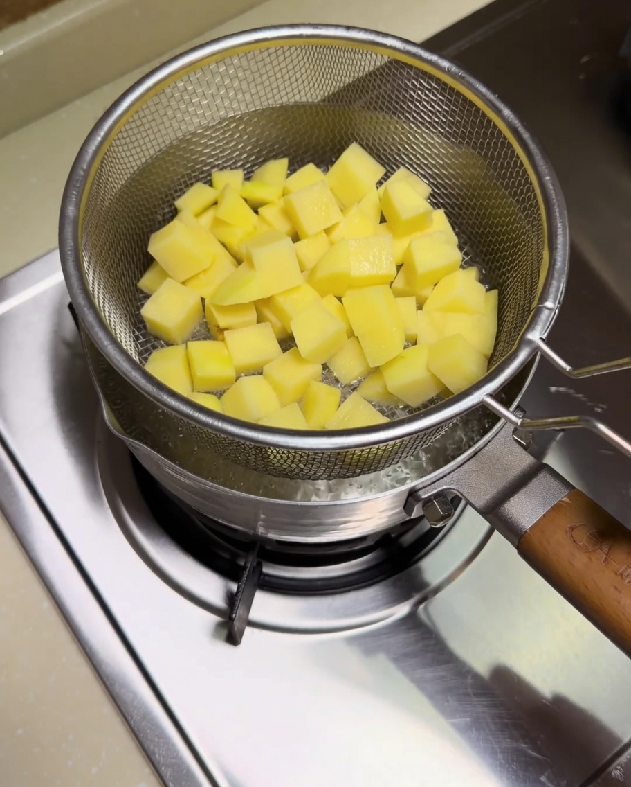 Patatas cortadas en dados cociéndose en un colador de metal dentro de una olla con agua hirviendo.