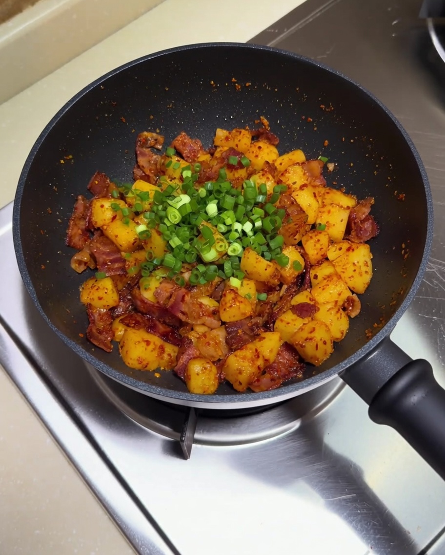 Una sartén llena de patatas y bacon crujiente, cubierta con un generoso puñado de cebolleta fresca picada lista para remover.