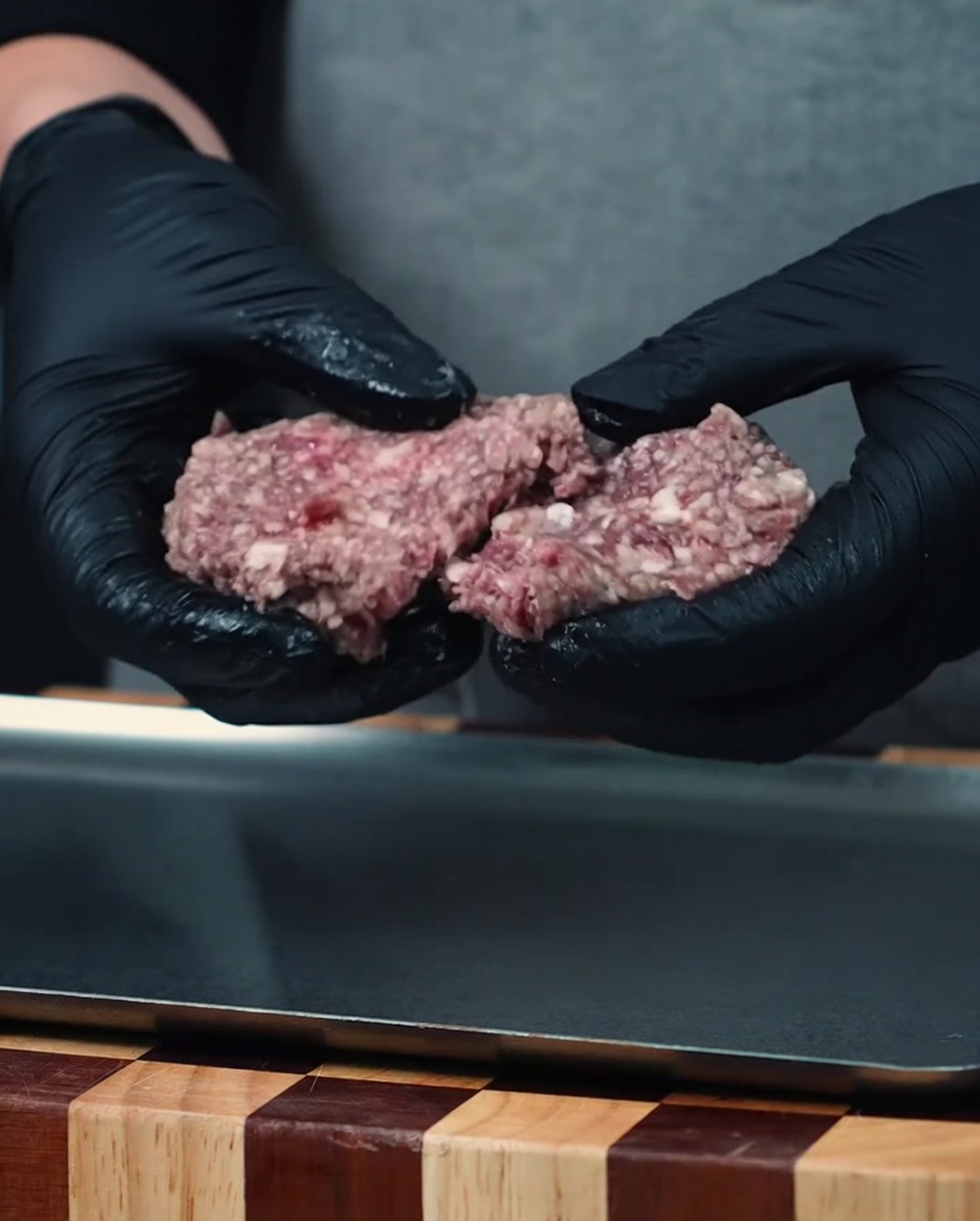 Dos bolas de carne picada de wagyu cruda siendo sostenidas con manos con guantes negros sobre una bandeja de metal.
