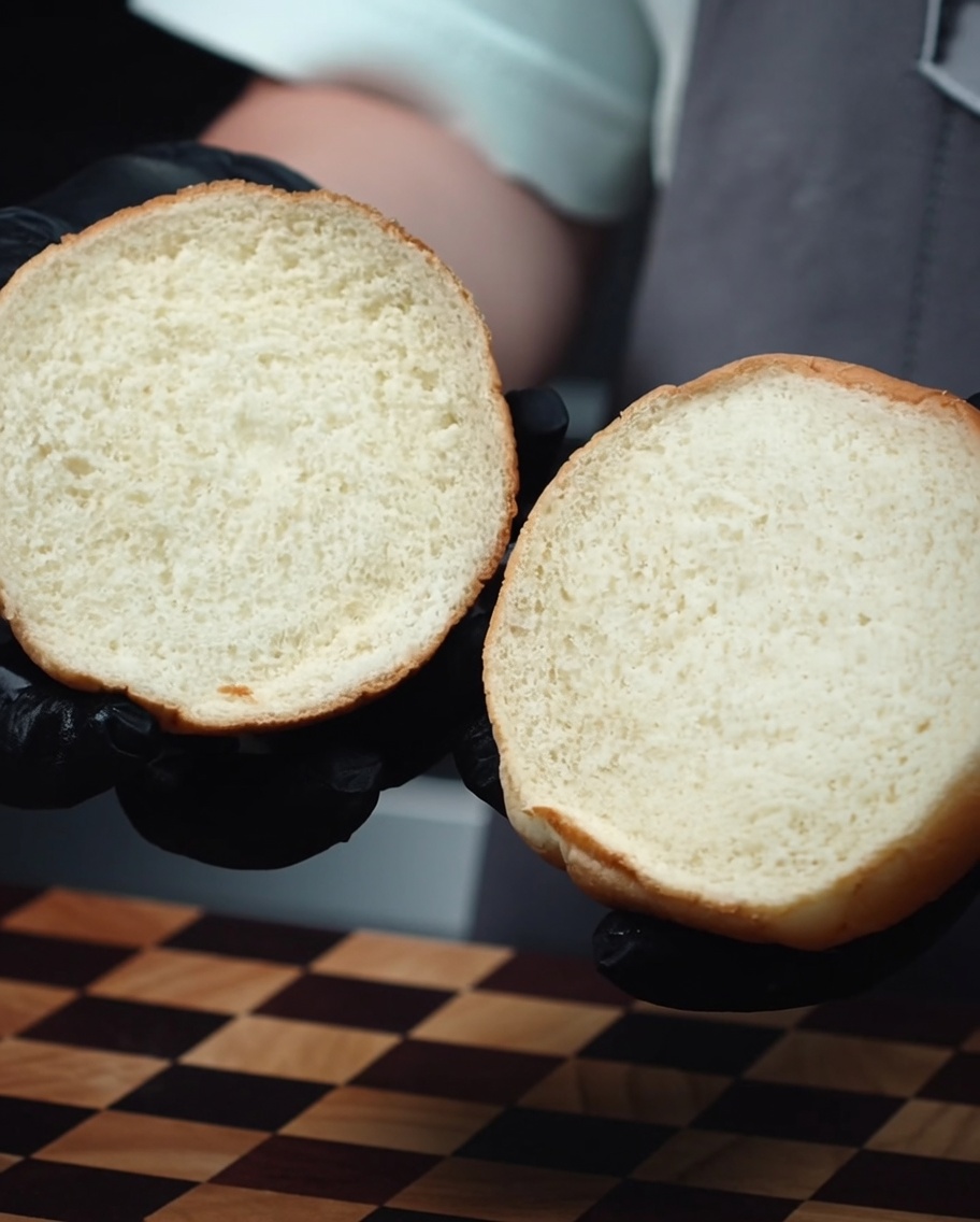 Dos mitades de un pan de hamburguesa suave, mostrando el interior tostado dorado, sostenidas por manos con guantes.