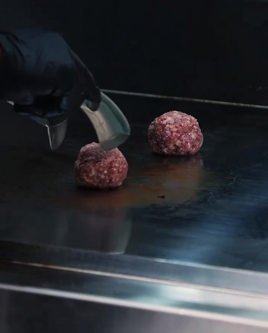 Un aplastador de metal pesado presionando una bola de carne cruda en una plancha caliente.