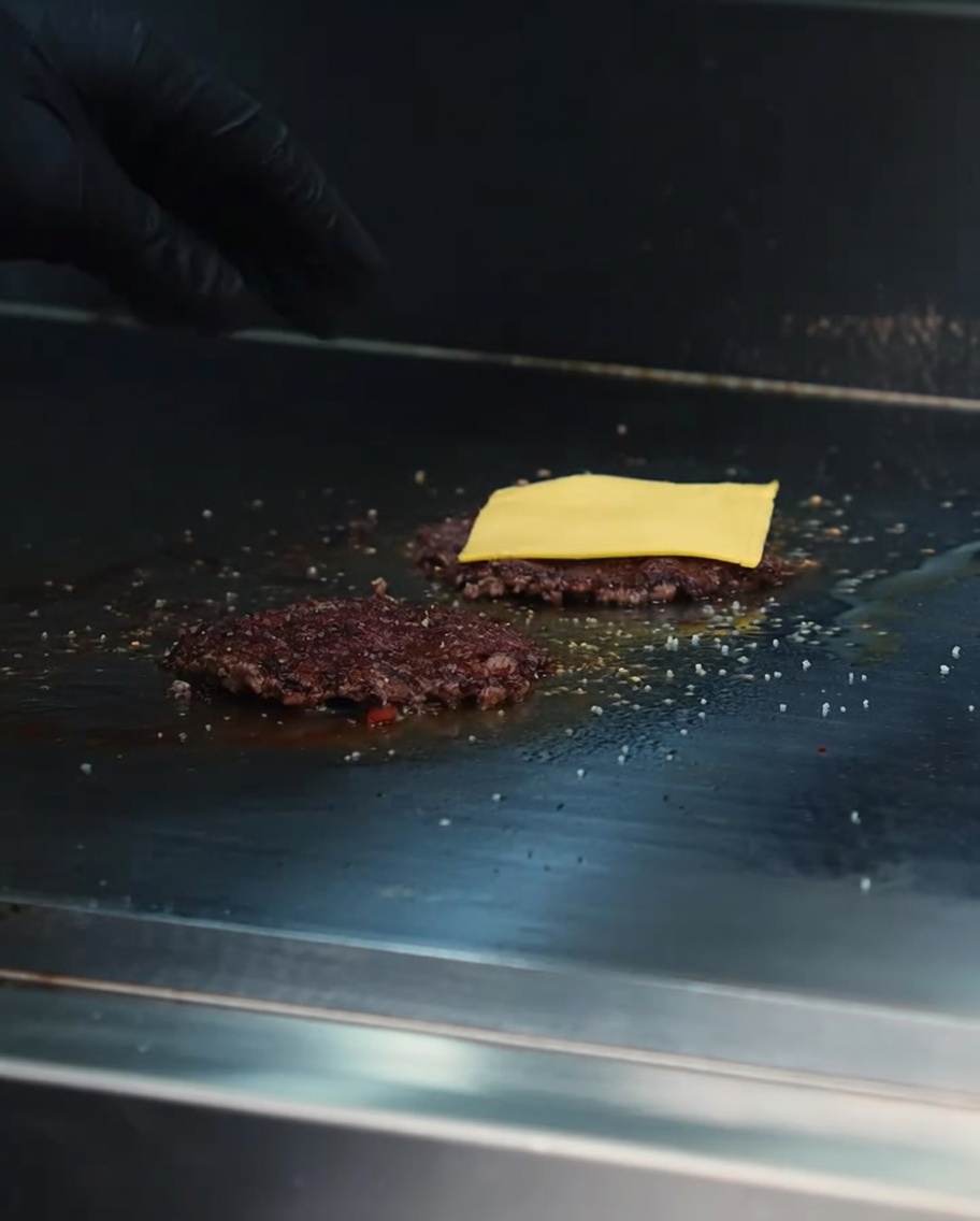 Lonchas de queso cheddar derritiéndose sobre hamburguesas de Wagyu en una plancha caliente.