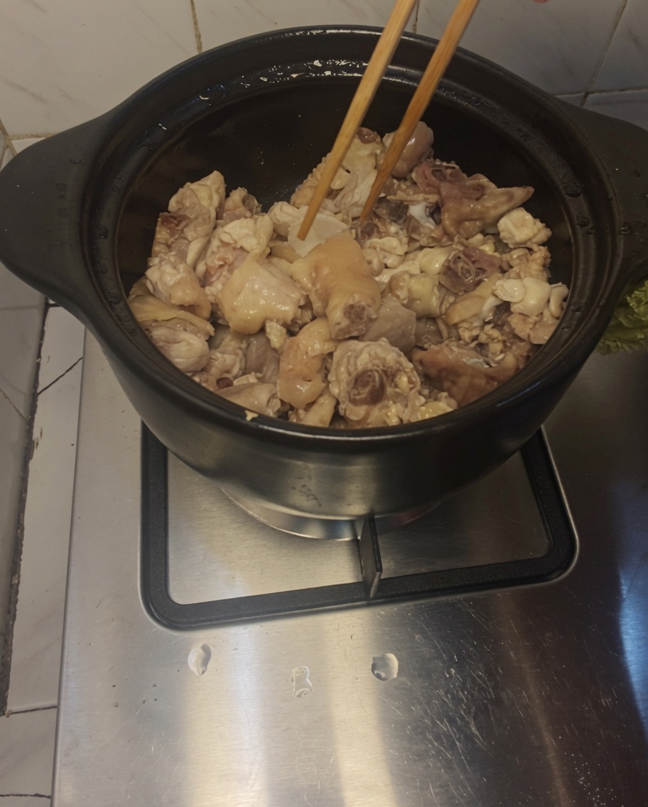 Palillos de madera removiendo trozos de pollo y rodajas de jengibre dentro de una olla de barro.