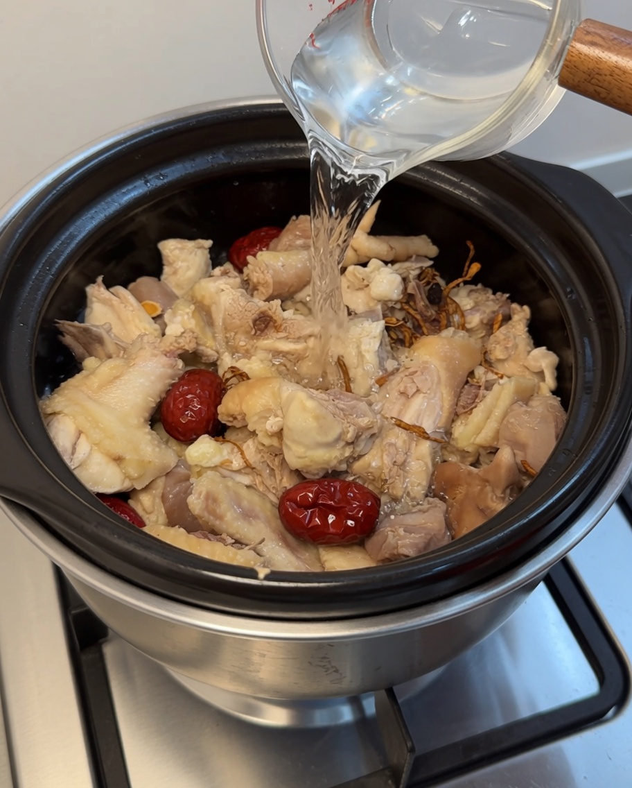 Agua clara vertiéndose desde un vaso medidor a una olla de barro con pollo y hierbas.