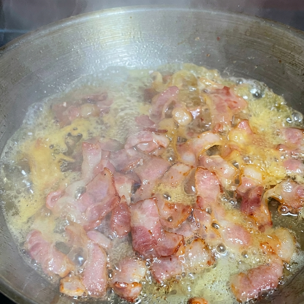 Espaguetis mezclándose en una sartén con bacon frito y un chorrito de agua de cocción.