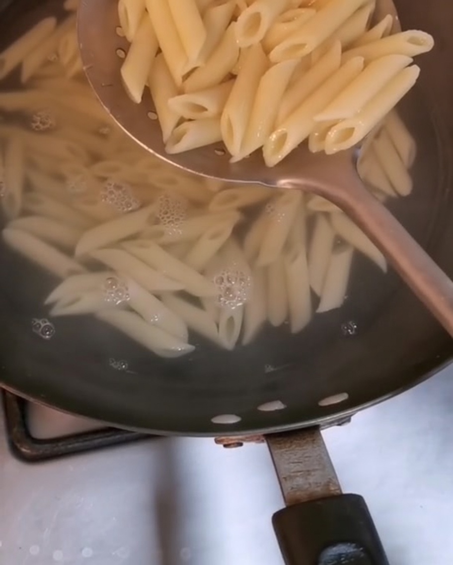 Pasta penne siendo retirada de agua hirviendo con una cuchara ranurada.