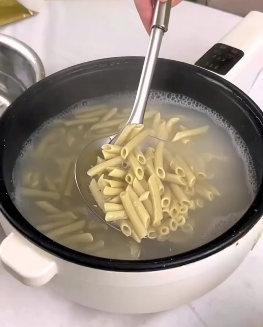 Pasta penne cocida siendo transferida de una olla a un recipiente con agua fría usando una espumadera.