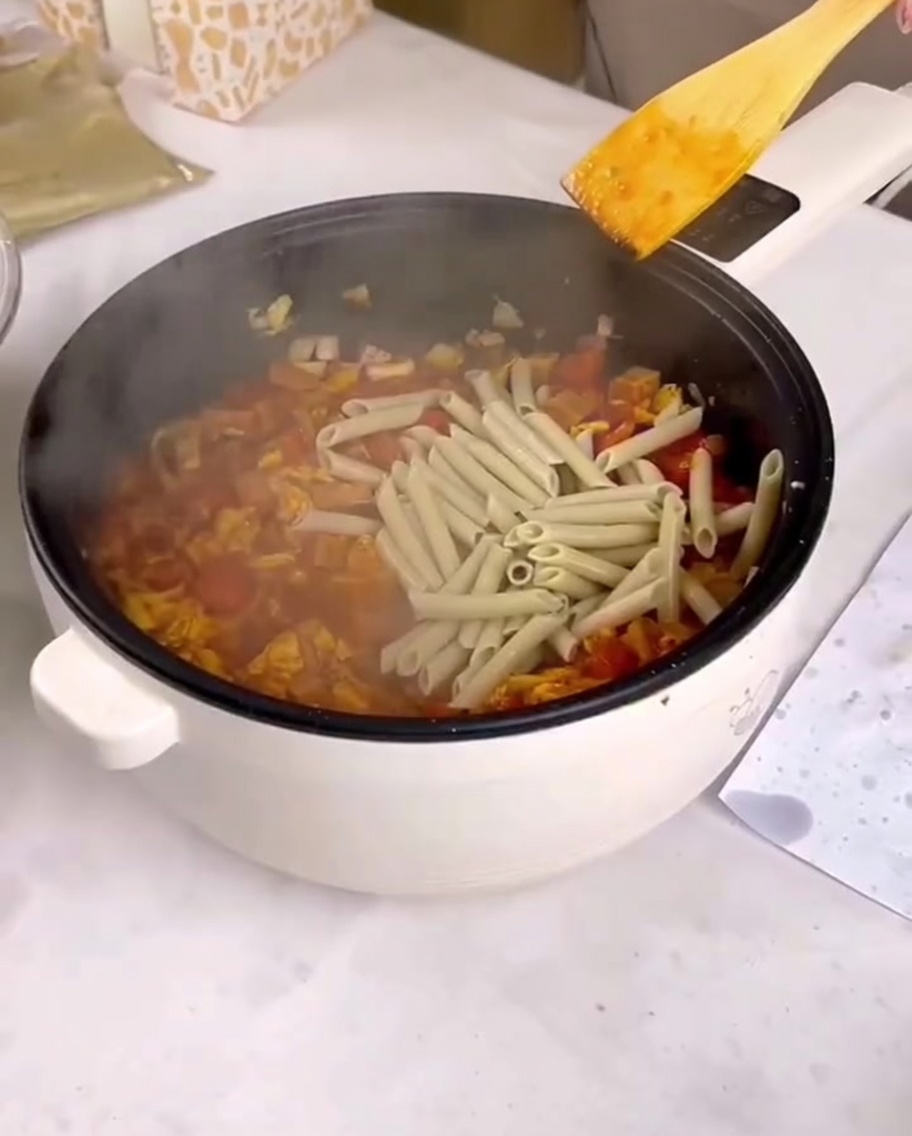 Penne cocido siendo añadido a una sartén con salsa e ingredientes salteados.