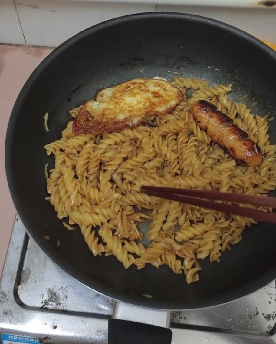 Huevo frito y salchicha servidos sobre una sartén de fusilli a la pimienta negra.