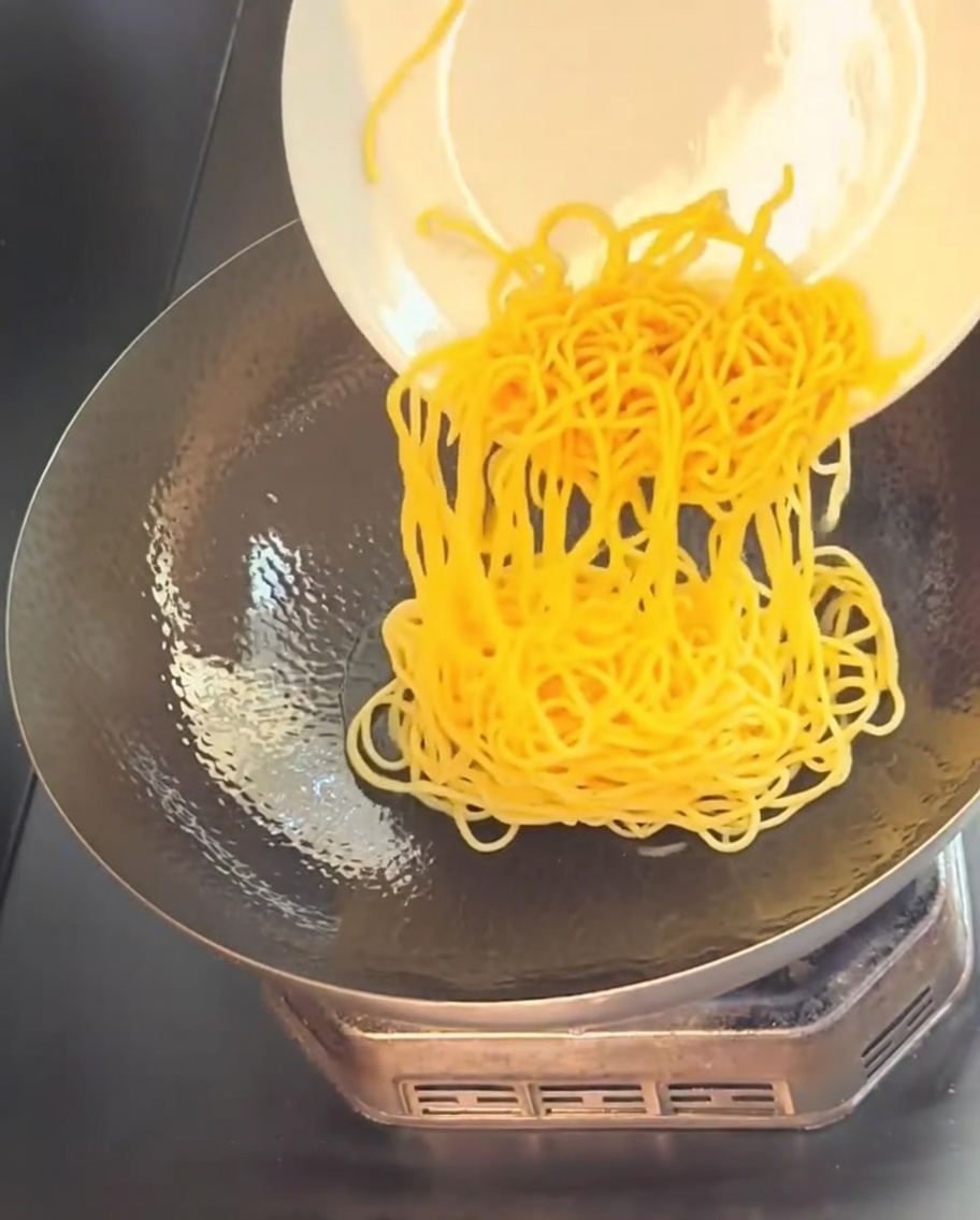 Espaguetis amarillos cocidos deslizándose desde un plato blanco hacia un wok caliente con aceite.