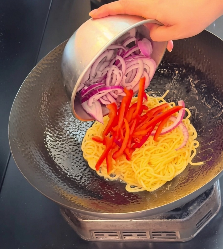 Vista en primer plano de tiras de ternera, pimiento rojo vibrante y cebolla morada siendo vertidos en un wok con espaguetis cocidos.