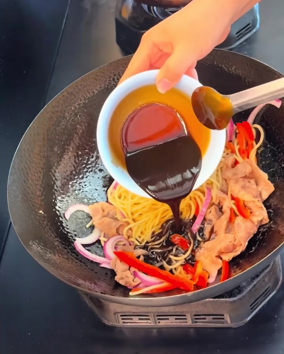 Una salsa oscura y rica de pimienta negra siendo vertida desde un pequeño cuenco blanco sobre los fideos, la carne y las verduras en un wok.