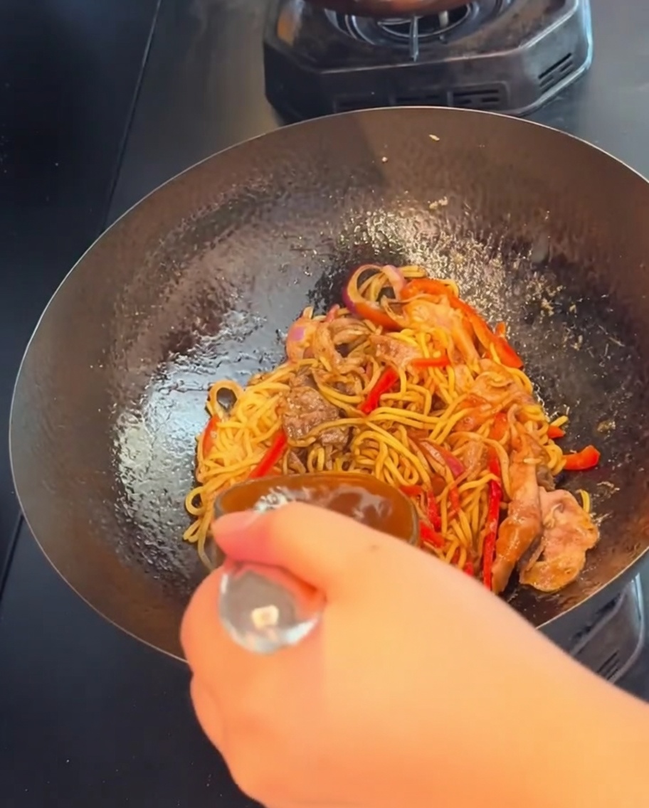 Una mano sosteniendo una cuchara, removiendo y salteando los espaguetis con ternera a la pimienta negra a fuego fuerte en un wok.