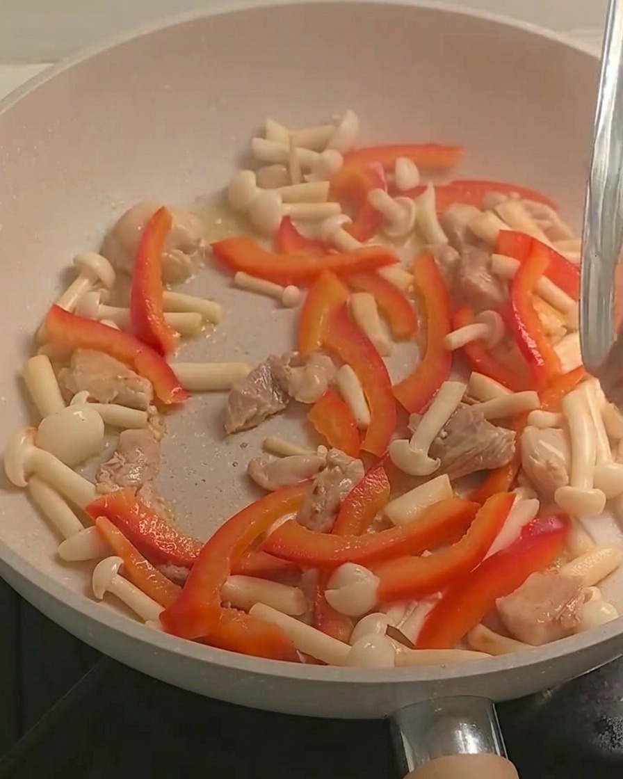 Pasta fusilli cocida siendo añadida a la sartén que contiene el pollo salteado y los vegetales coloridos.