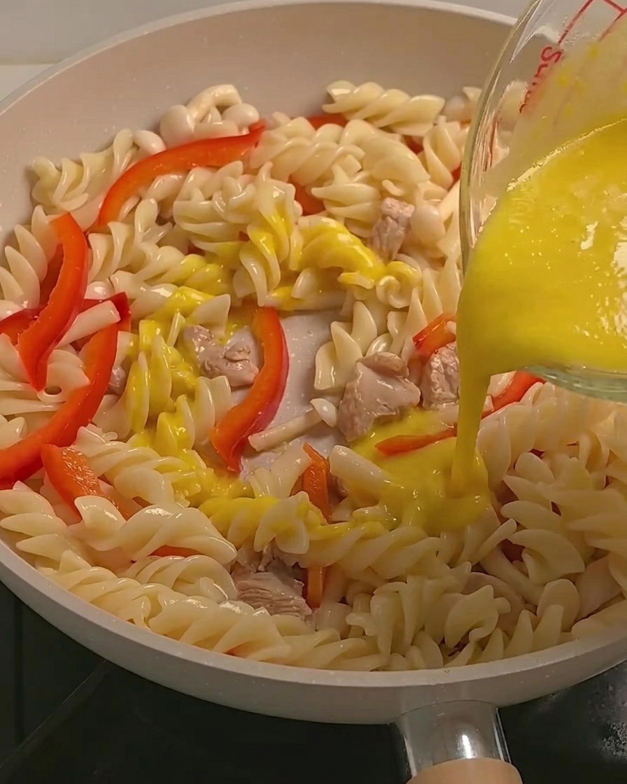 Salsa cremosa de calabaza amarilla siendo vertida sobre la pasta fusilli, el pollo y los vegetales en una sartén.