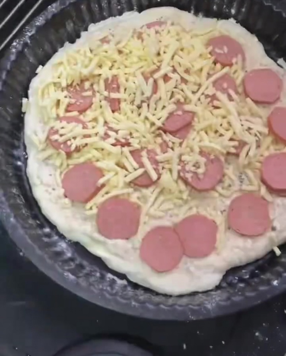 Una capa final y generosa de queso mozzarella rallado siendo esparcida sobre la pizza con salchichas.