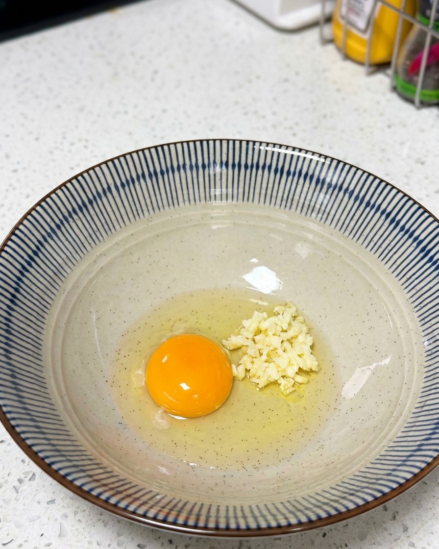 Un bol que contiene una yema de huevo cruda, ajo picado y una porción de mayonesa.