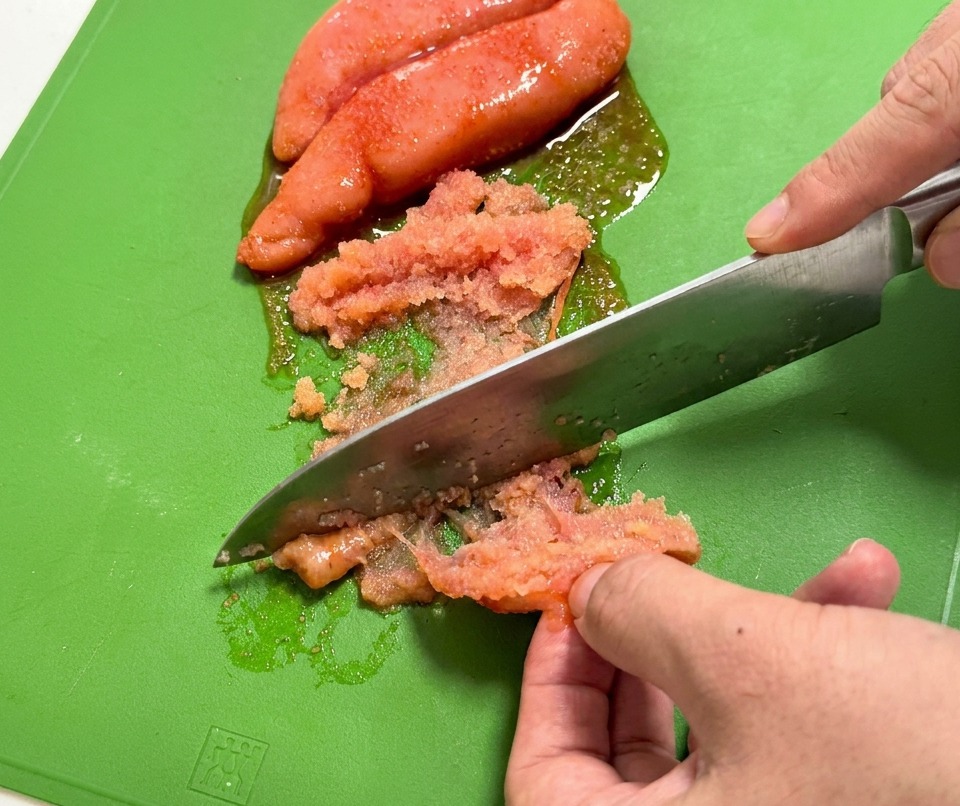 Un cuchillo raspando la delicada hueva de pescado rosada de su saco sobre una tabla de cortar.