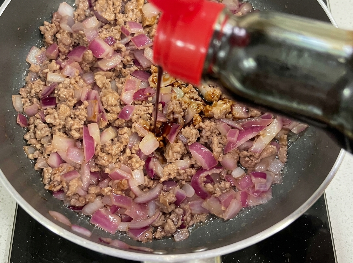 Salsa de soja oscura y kétchup siendo añadidos a la sartén con carne y cebolla.