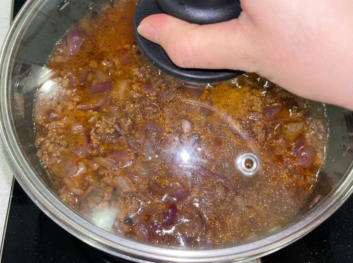 Mezcla de carne y cebolla en una sartén tapada con una tapa de cristal tras añadir agua.
