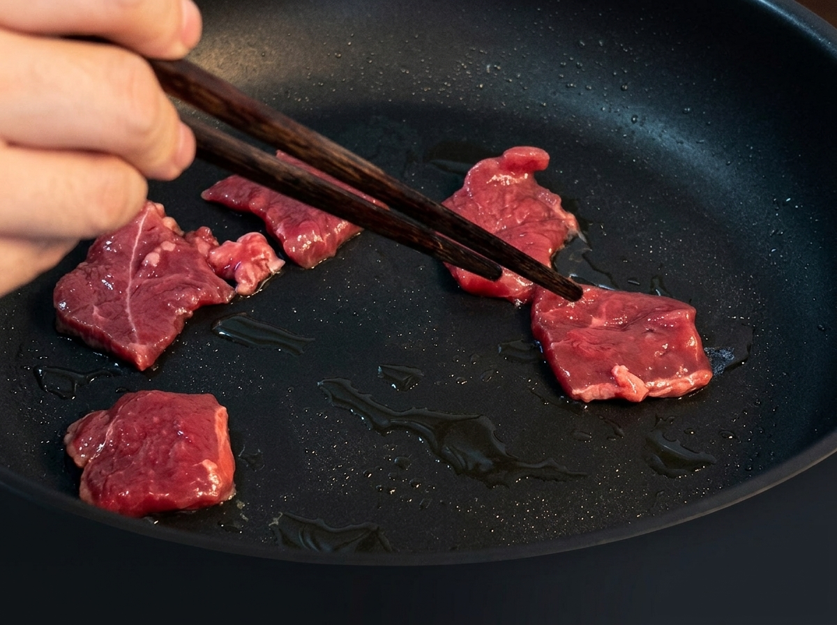 Tiras finas de ternera salteándose en una sartén caliente con aceite, dorándose a medida que se cocinan.