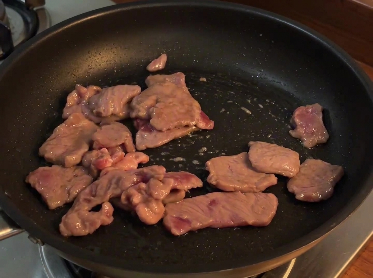 Rodajas de cebolla roja añadidas a la sartén con la ternera cocinada, siendo mezcladas a fuego.