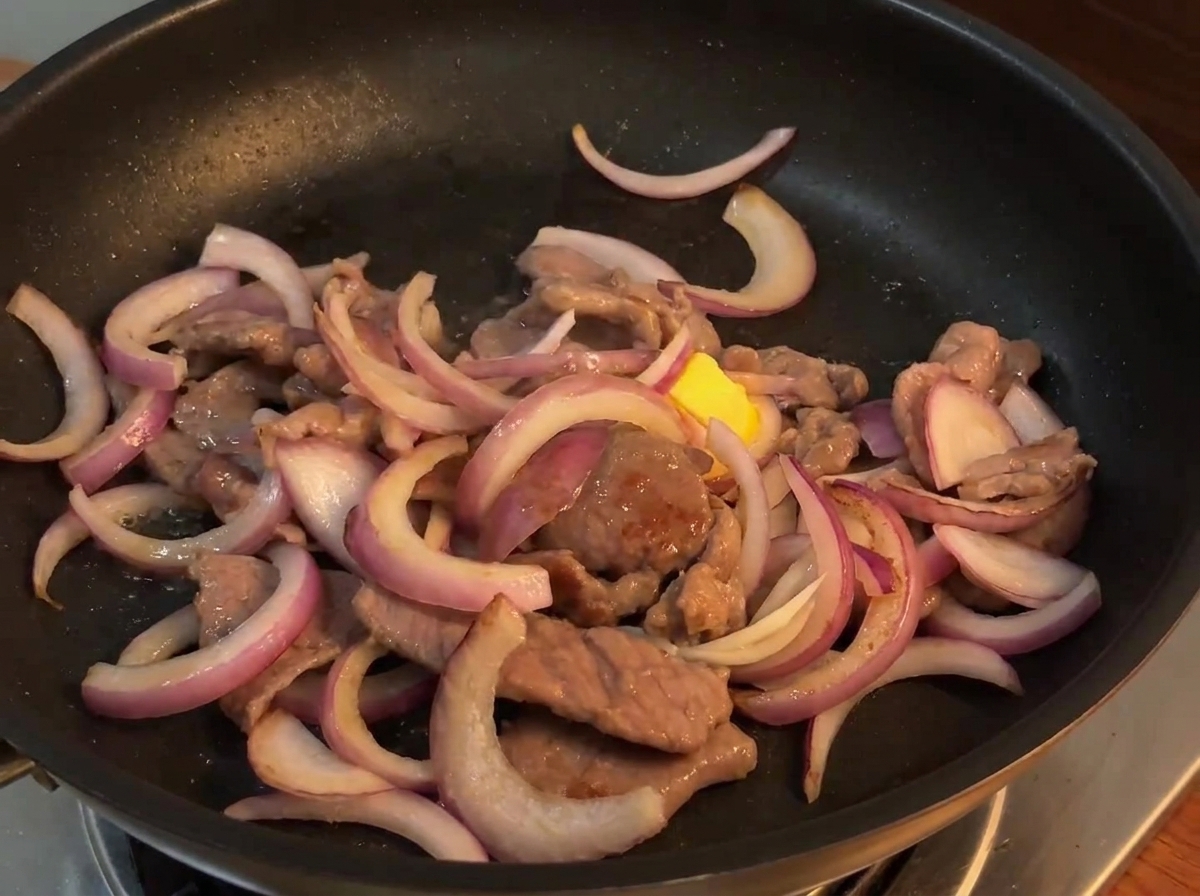 Salsa de salteado siendo vertida desde un tazón pequeño sobre la mezcla de ternera y cebolla en una sartén caliente.