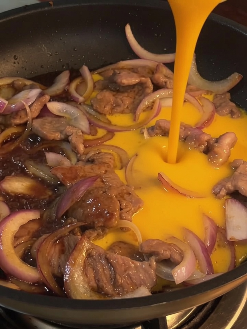 Primer plano de huevos batidos siendo vertidos en una sartén sobre la ternera y cebollas salteadas.