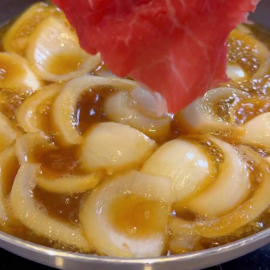 Láminas finas de ternera cruda siendo colocadas en una sartén con cebollas y salsa.