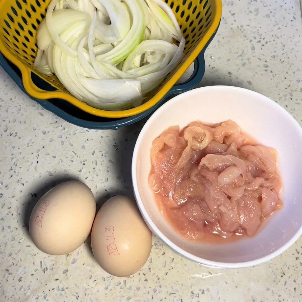 Trozos de pechuga de pollo cruda marinando en un bol blanco junto a huevos frescos y cebollas cortadas.