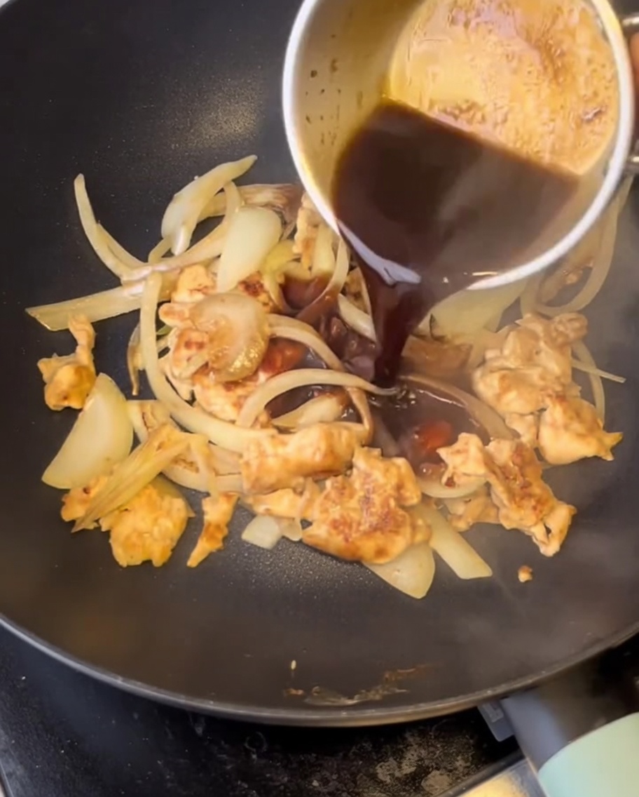 Vertiendo salsa oscura sabrosa desde una pequeña olla sobre trozos de pollo cocinado y cebollas cortadas en una sartén caliente.