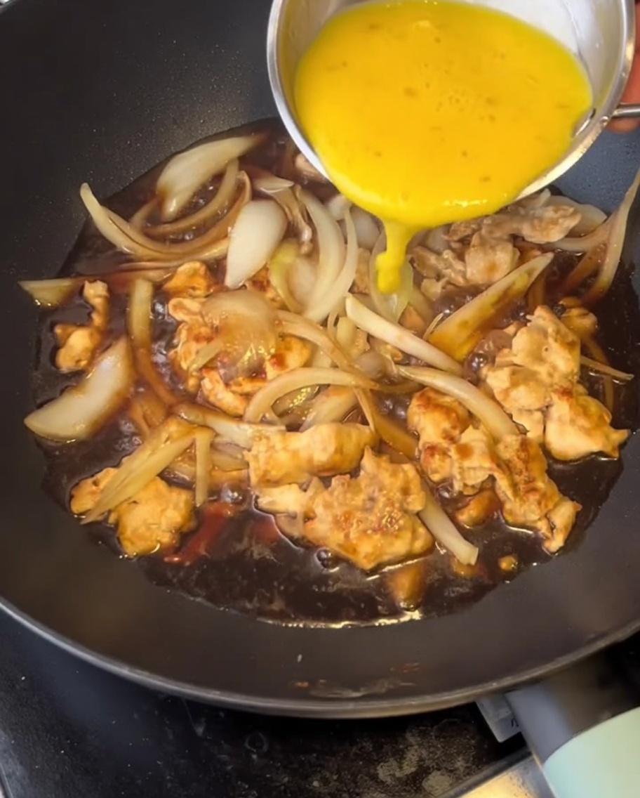 Vertiendo huevos batidos amarillos brillantes en una sartén llena de pollo, cebollas y salsa oscura sabrosa a fuego lento.