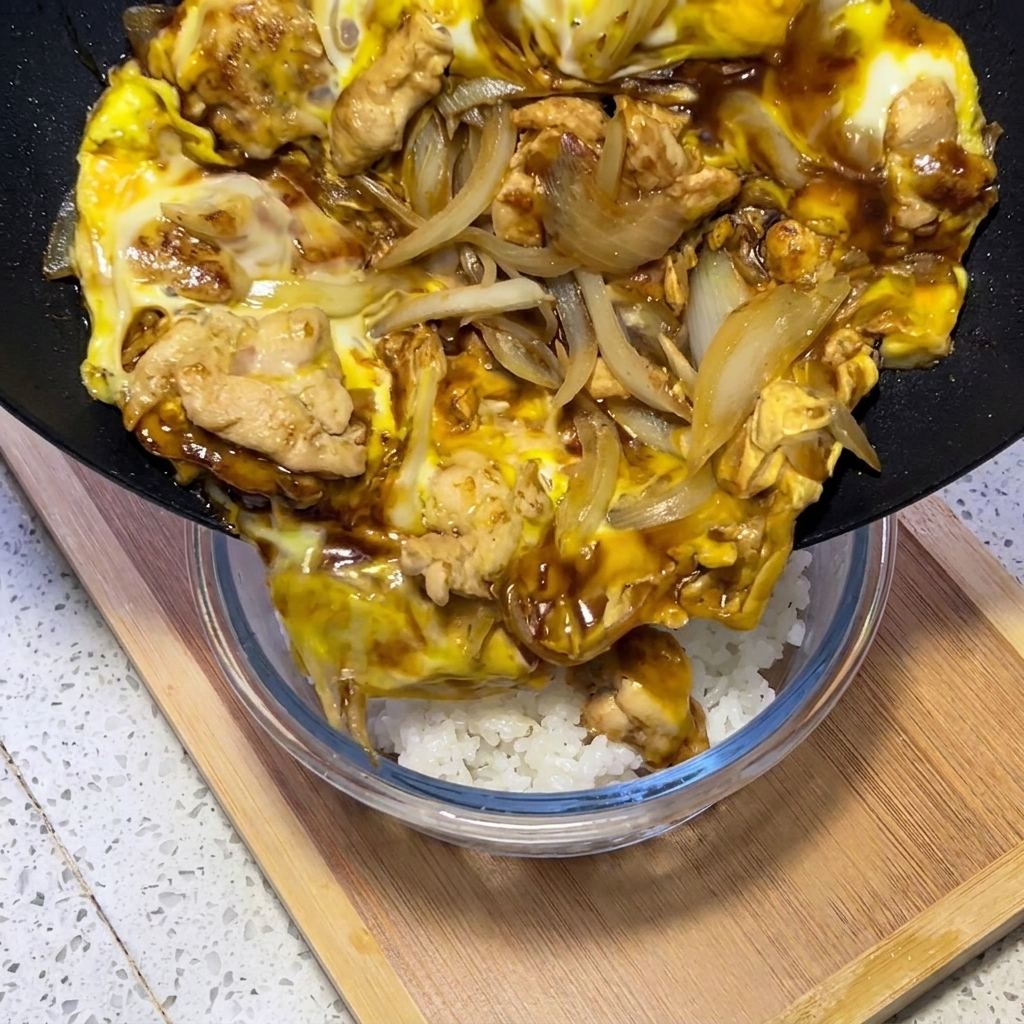 Una mezcla cocinada de pollo, huevo y cebollas en salsa sabrosa deslizándose desde una sartén negra directamente sobre un bol de arroz blanco.