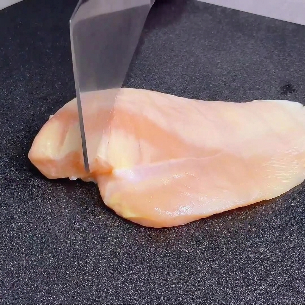 Aplanando una pechuga de pollo cruda con el borde posterior de un cuchillo grande sobre una tabla de cortar oscura.