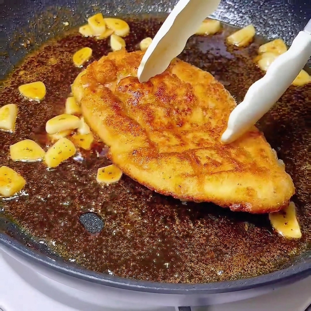 Pinzas colocando una pechuga de pollo dorada en un glaseado oscuro burbujeante con ajo laminado.