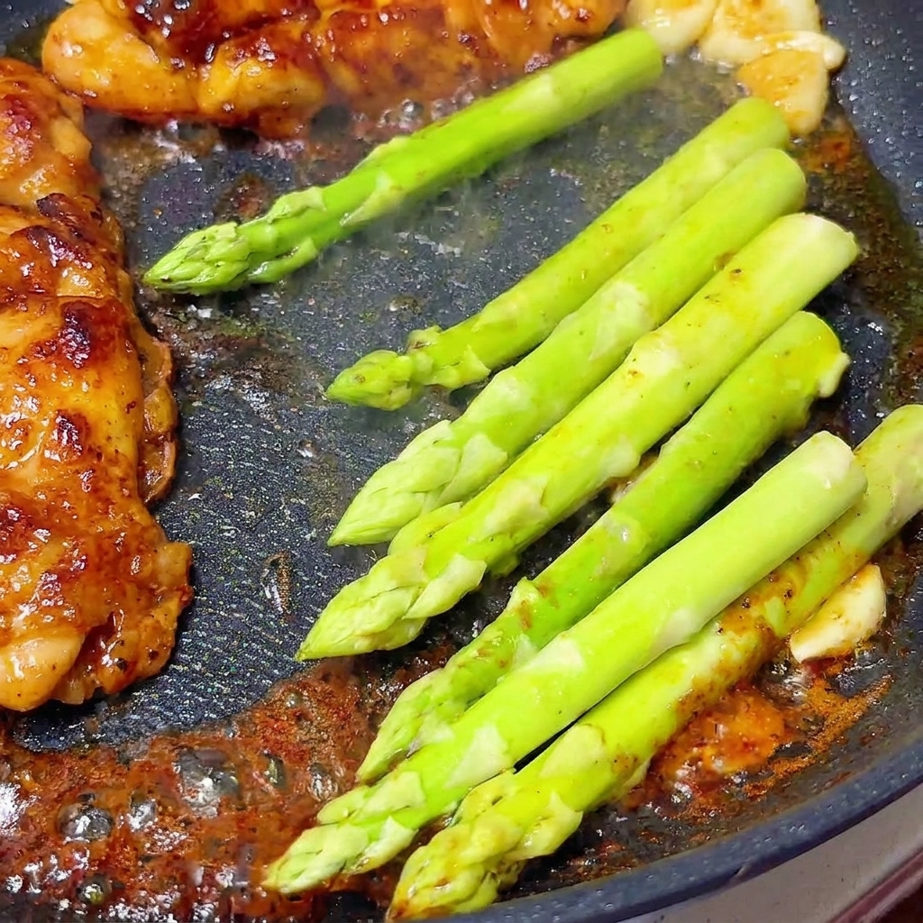 Tallos de espárragos frescos verde brillante cocinándose junto a pechugas de pollo glaseadas en una sartén.