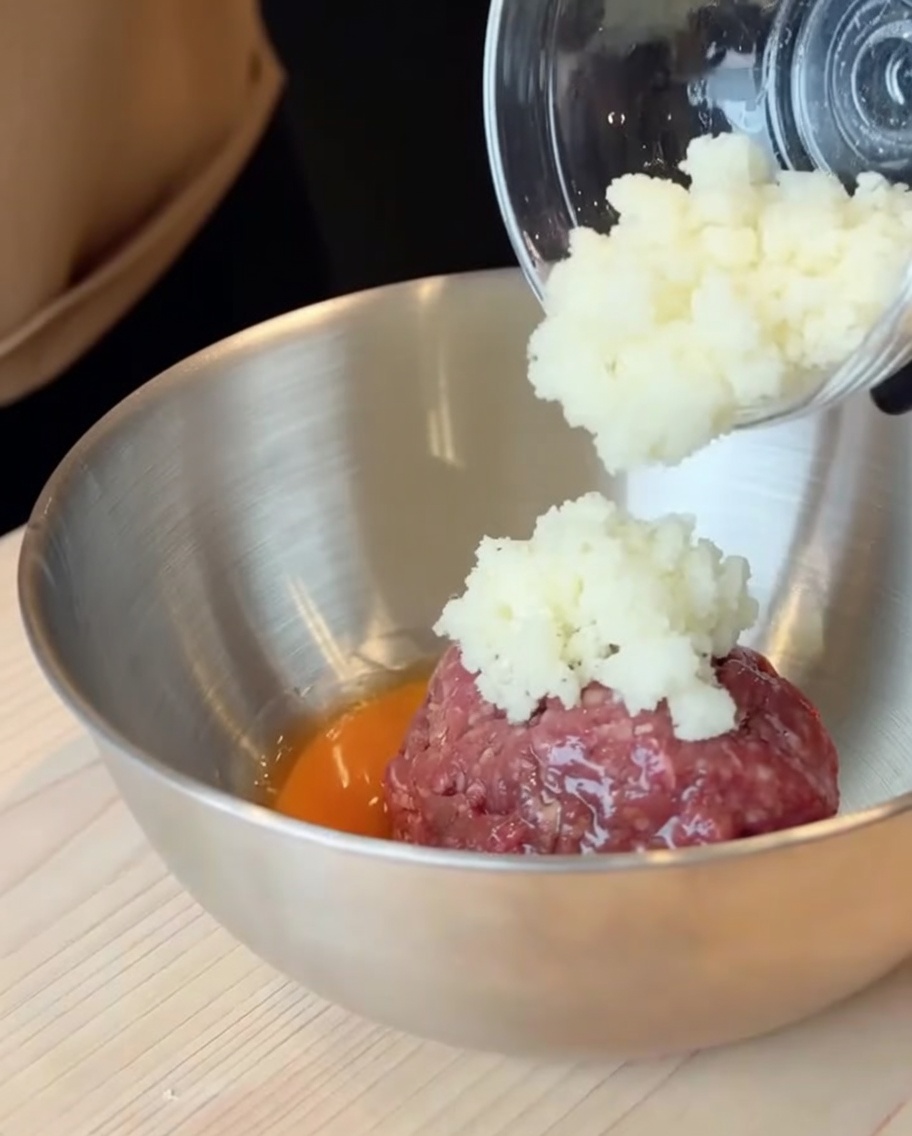 Carne picada cruda y un huevo roto en un cuenco grande de acero inoxidable con cebollas picadas siendo añadidas.