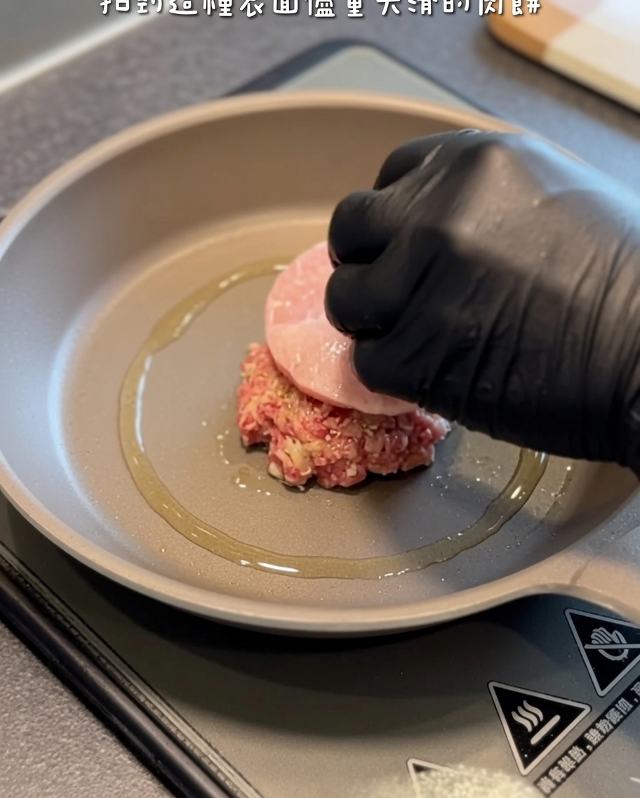 Una hamburguesa cruda siendo colocada con cuidado en una sartén caliente con aceite usando una mano enguantada y una espátula.