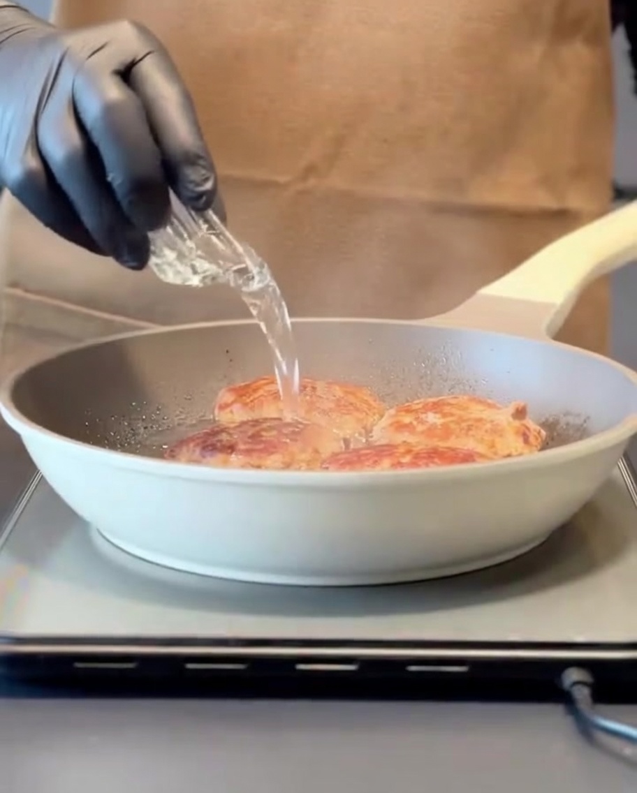 Agua siendo vertida en una sartén caliente con hamburguesas selladas, creando vapor antes de tapar con una tapa.