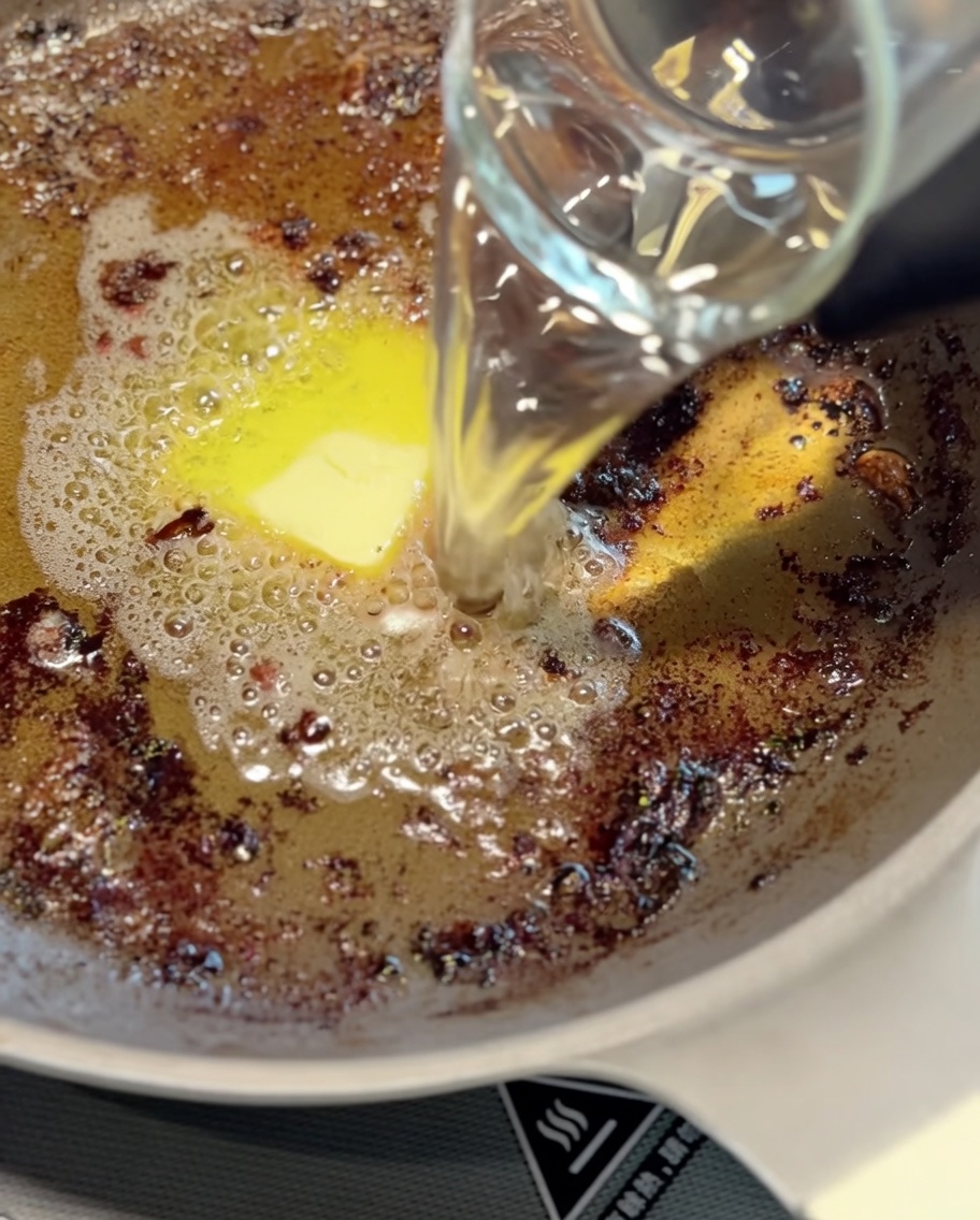 Un cucharón de metal removiendo los jugos de la sartén con agua para formar una base de salsa.