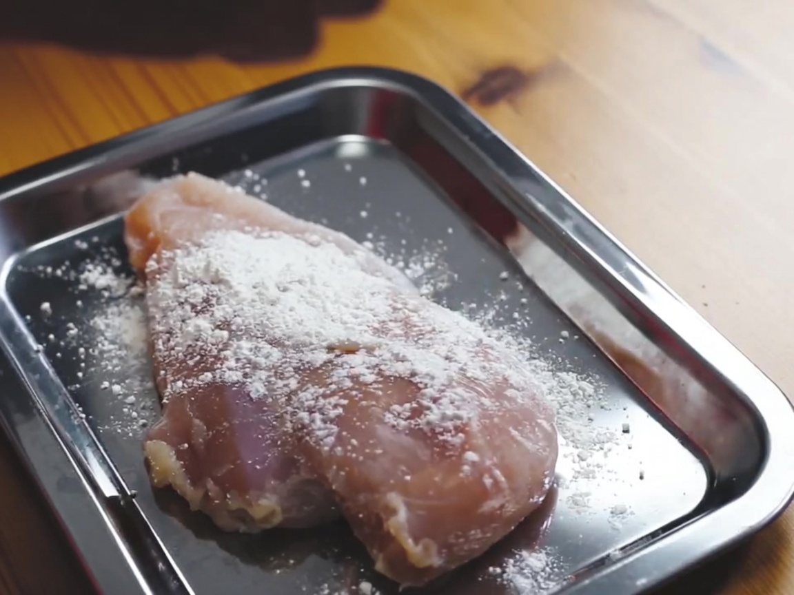 Pechuga de pollo cruda siendo cubierta de harina en una bandeja metálica.