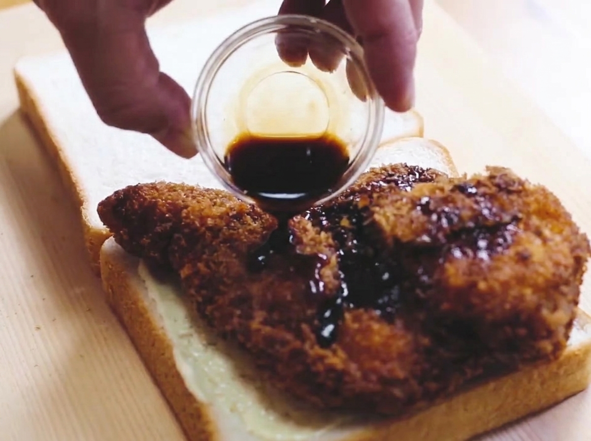 Un filete de pollo frito dorado siendo rociado con salsa oscura mientras descansa sobre una pieza de pan blanco.