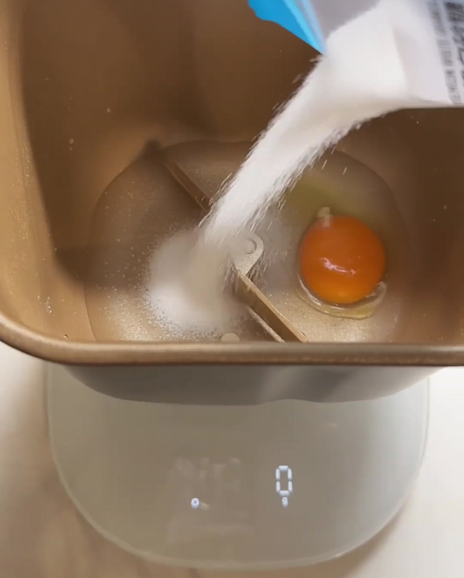 Ingredientes para la masa de pan, incluyendo un huevo y azúcar, siendo añadidos al recipiente de la panificadora.