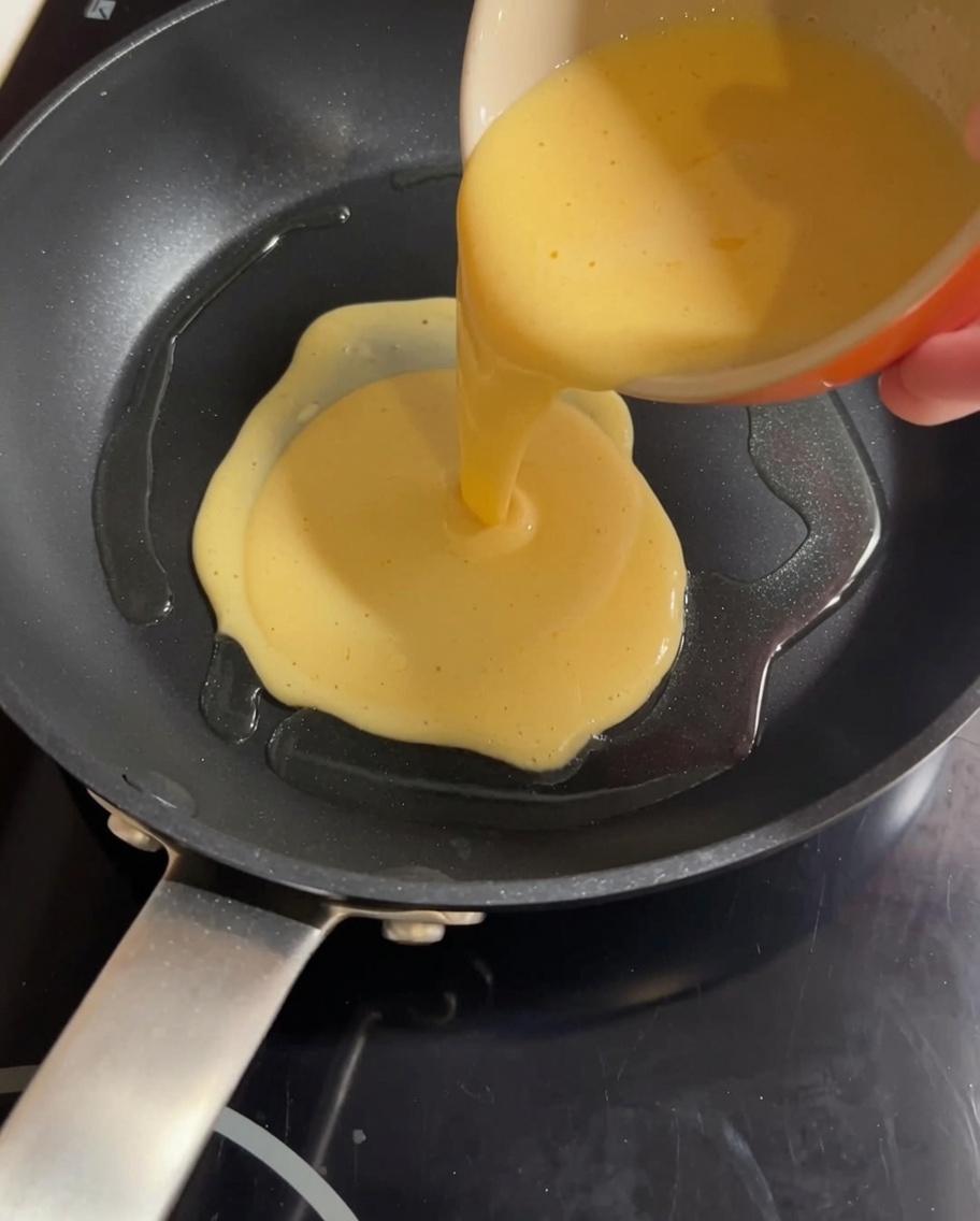Mezcla amarilla de huevo y leche vertiéndose en una sartén caliente, lista para ser revuelta.