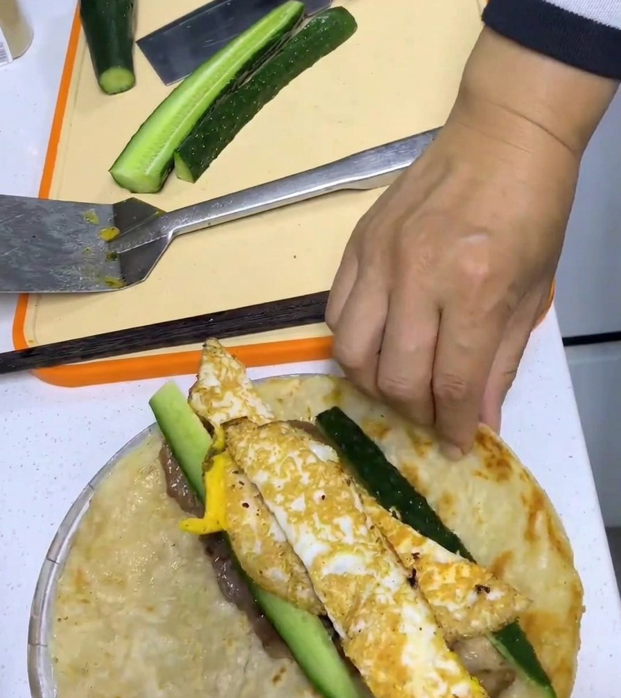 Una mano enrollando fuertemente el pancake alrededor del cerdo, huevo y pepino.