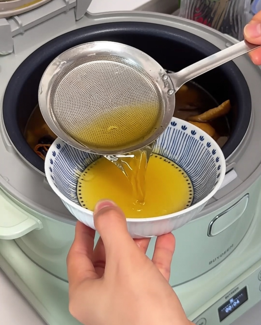 Primer plano de un colador de malla fina retirando la grasa de pollo clara y dorada de la parte superior de la sopa a fuego lento en una olla eléctrica.