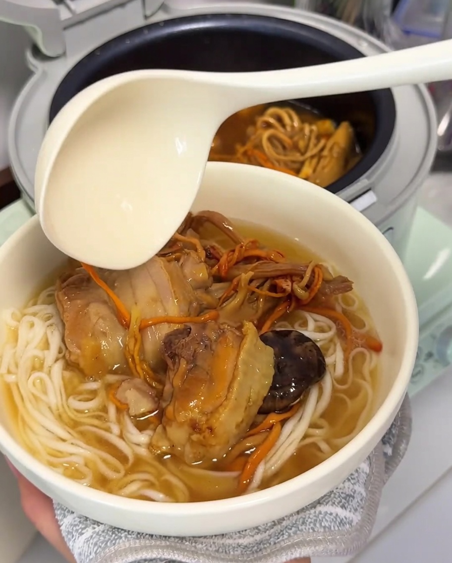 Un cucharón grande vertiendo la rica y dorada sopa de pollo y setas sobre un tazón de fideos recién cocidos.